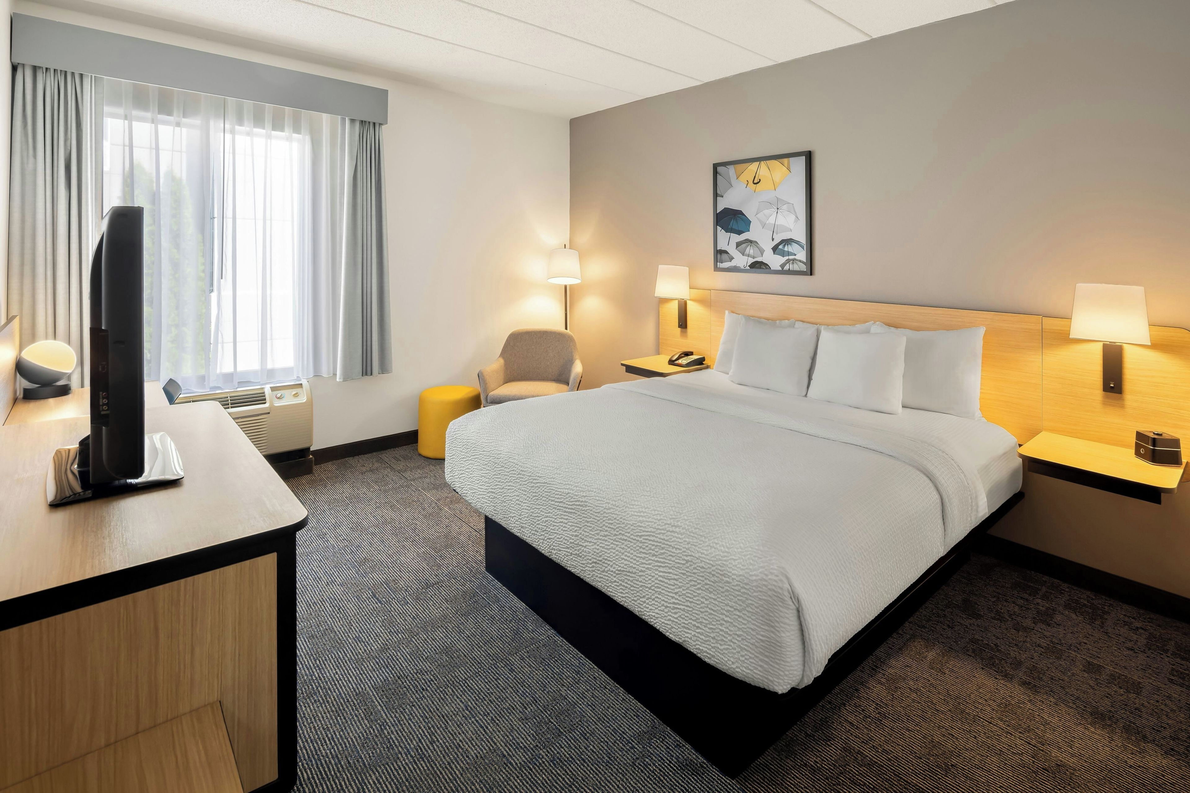 A hotel room with a bed, a desk with a television, two bedside lamps, a chair, and a wall-mounted artwork.