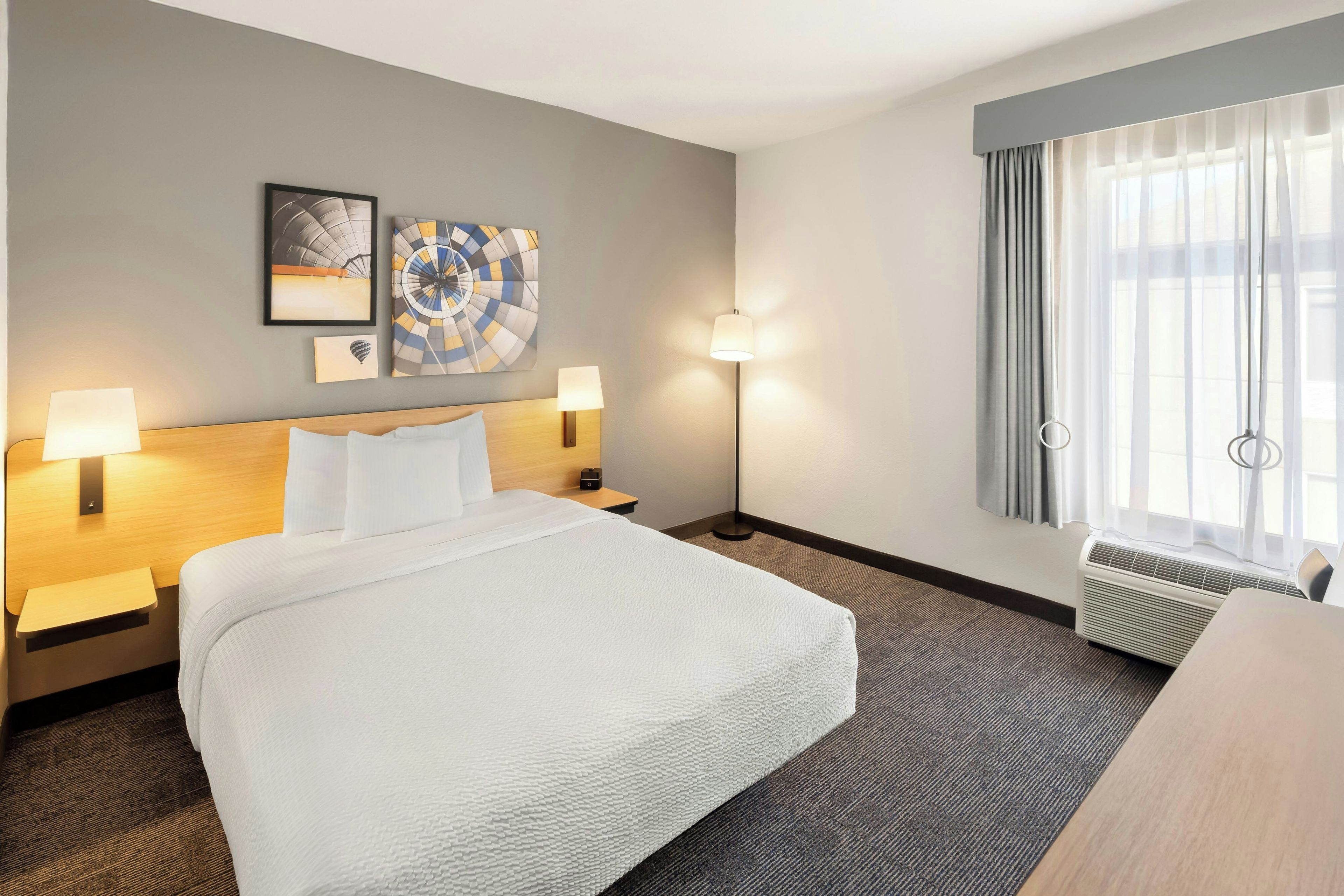 A hotel room with a bed, two bedside lamps, a floor lamp, a window with curtains, and two wall art pieces.