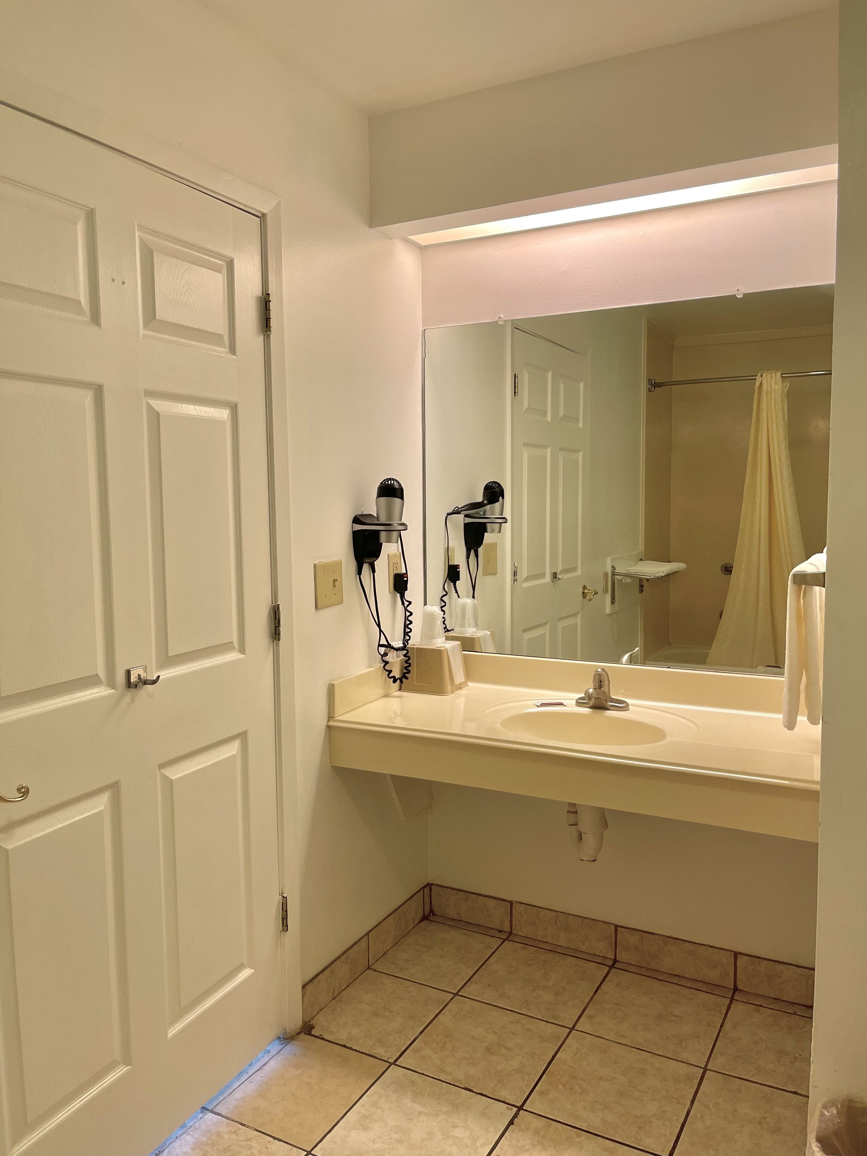 Casa de banho com lavatório branco, dois secadores de cabelo fixados na parede, espelho sobre o lavatório e cortina de chuveiro visível no reflexo.