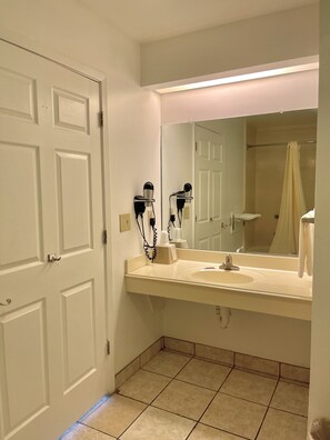 A bathroom with a white sink, two hair dryers mounted on the wall, a mirror above the sink, and a shower curtain visible in the reflection.