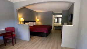 A compact hotel room with a bed, a desk, and a chair. There is a small table with a lamp, and a TV mounted on the wall. - Majestic Inn (Longview)