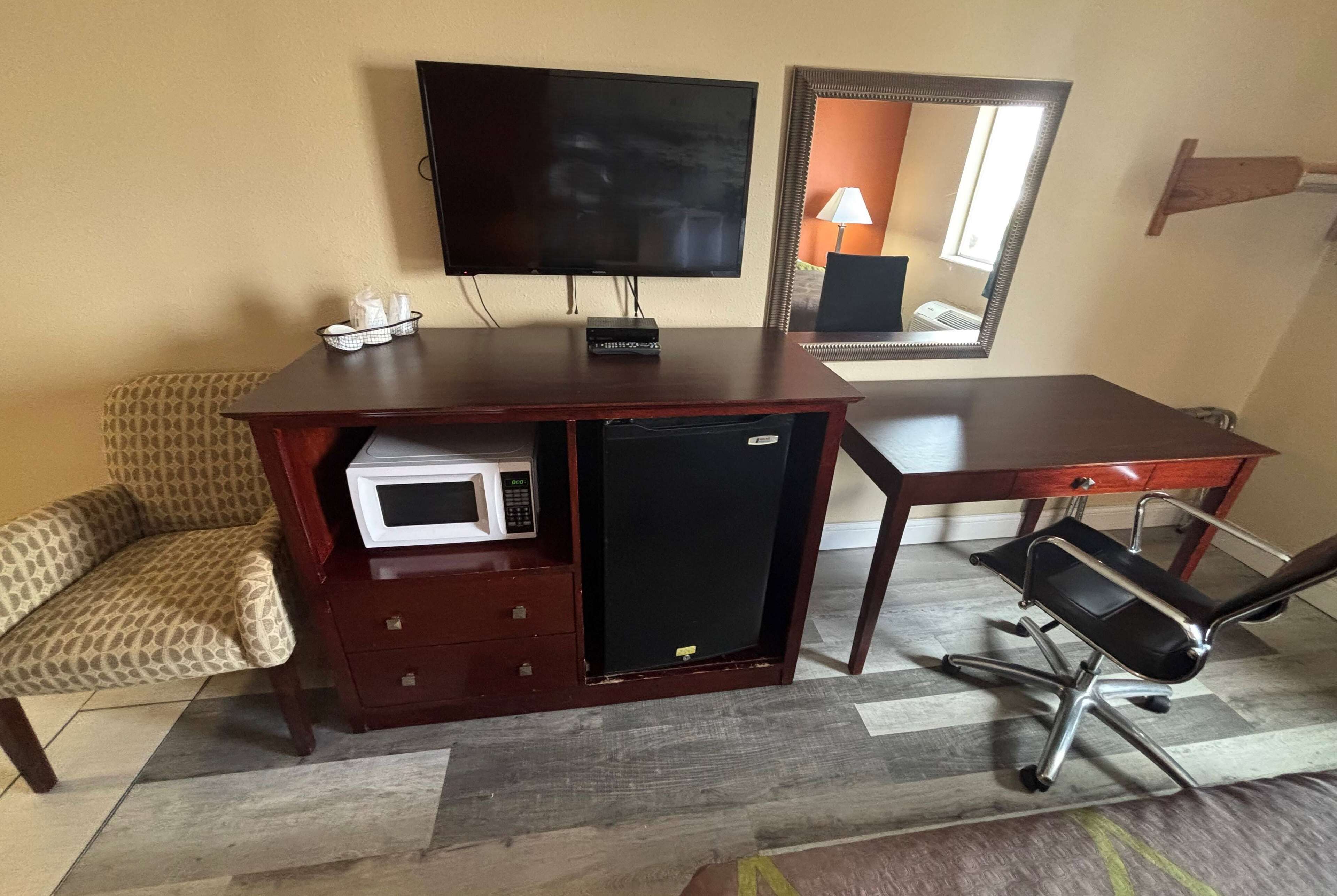 A compact hotel room with a wooden desk, a television, a microwave, a small refrigerator, a patterned armchair, and a black office chair.