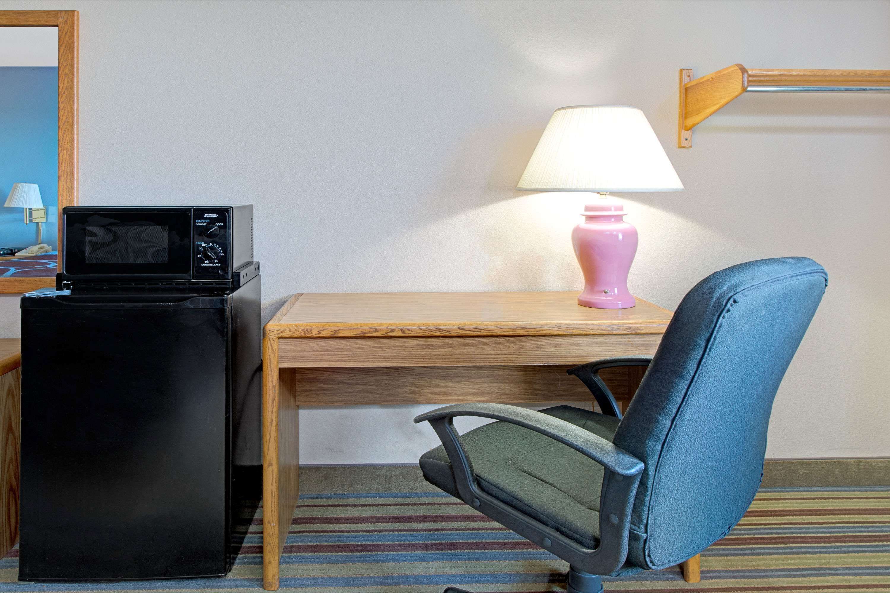 Una habitación de hotel con un escritorio de madera, un refrigerador negro, una silla azul, una lámpara rosa y una alfombra a rayas.