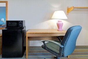 Una habitación de hotel con un escritorio de madera, un refrigerador negro, una silla azul, una lámpara rosa y una alfombra a rayas.