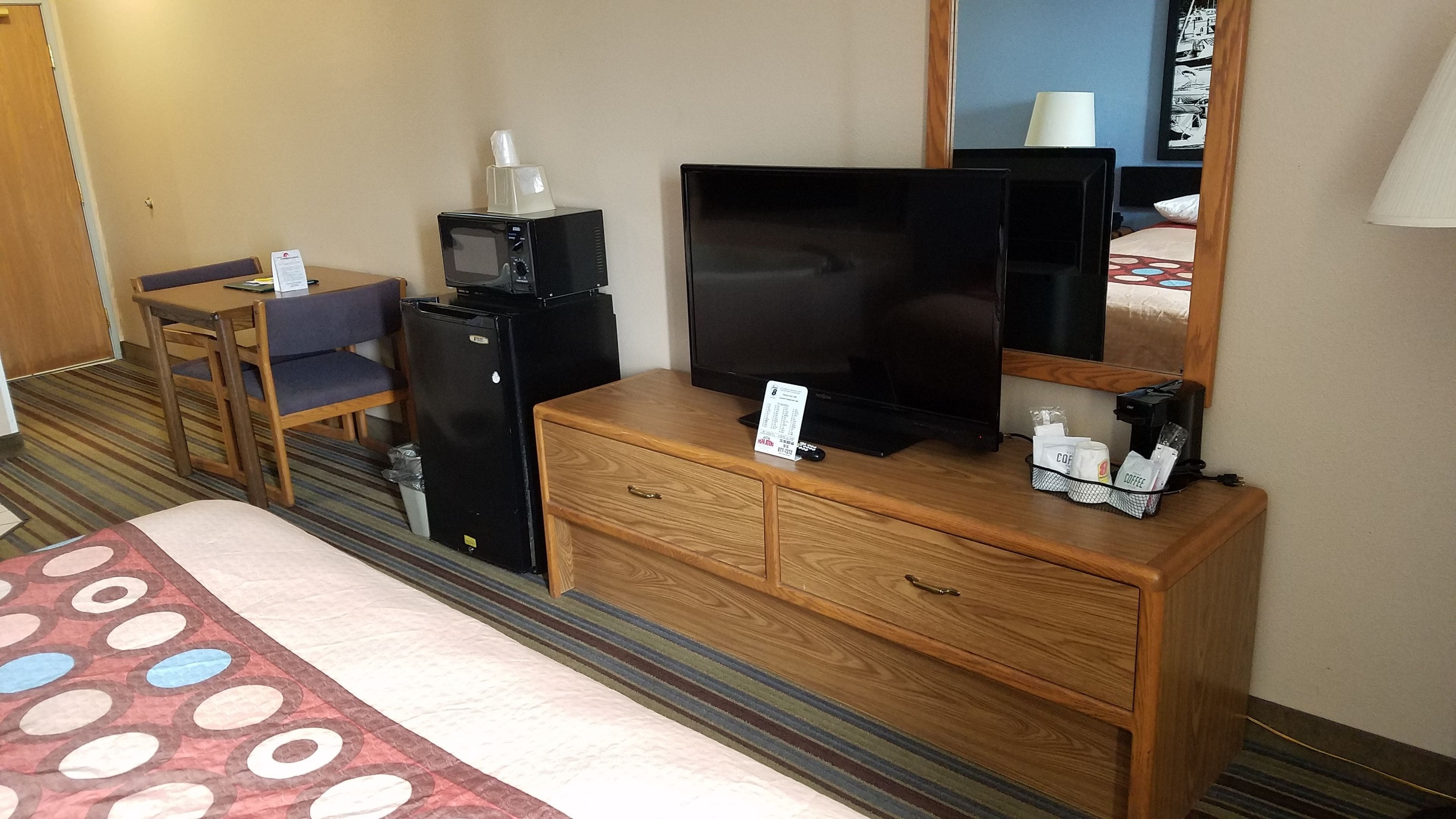 A hotel room with a bed, a wooden dresser, a television, a microwave, a refrigerator, a coffee maker, and a small table with a chair.