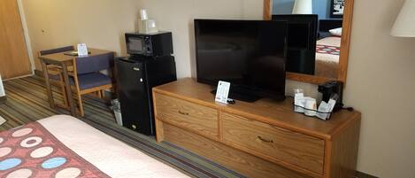 A hotel room with a bed, a wooden dresser, a television, a microwave, a refrigerator, a coffee maker, and a small table with a chair.