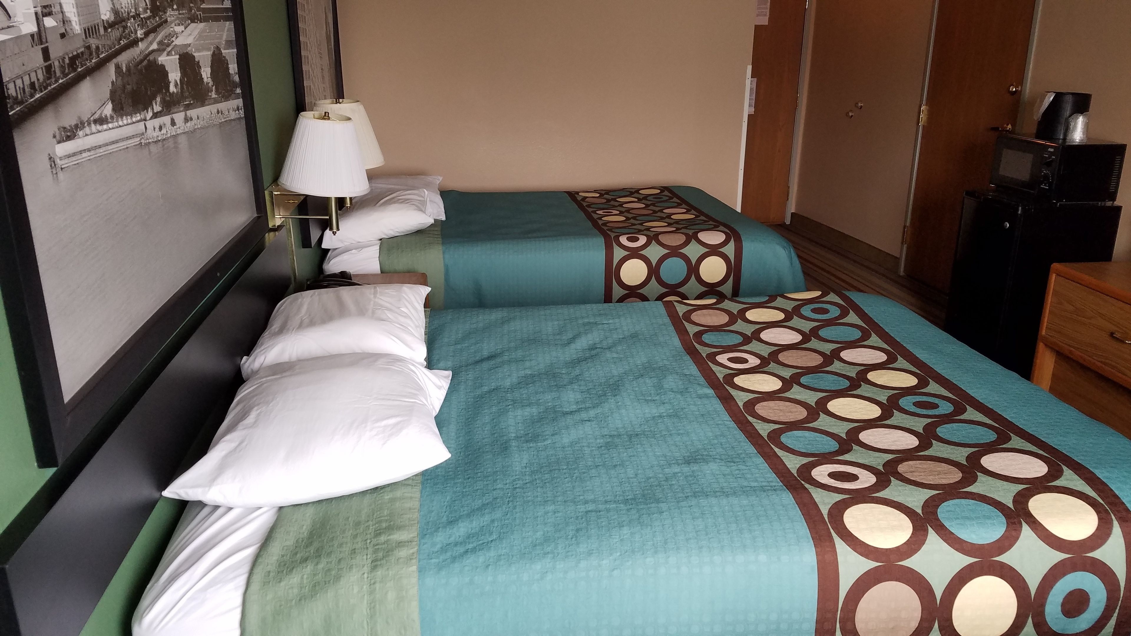 A hotel room with two beds, each with a teal bedspread and white pillows. There is a nightstand with a lamp, a wooden dresser, and a microwave on top.