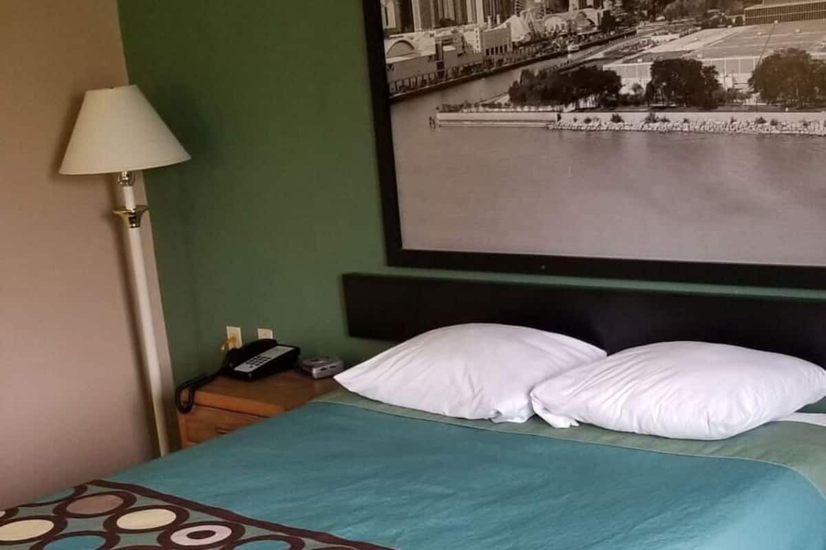 A hotel room with a bed, a cityscape artwork on the wall, a lamp, and a telephone on the nightstand.