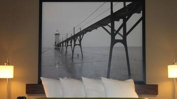 A hotel room with a large framed picture of a pier and lighthouse, a bed with white pillows, and two bedside lamps.