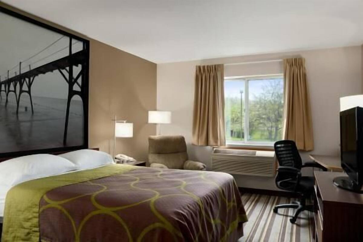 A hotel room with a bed, a desk, a chair, a TV, a window with curtains, and a painting on the wall.