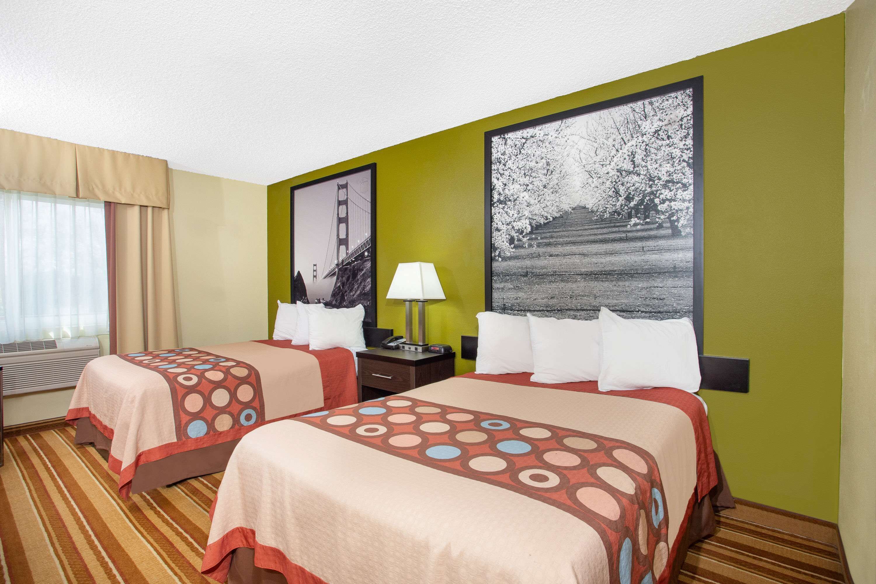 A hotel room with two beds, a window with curtains, a lamp, and artwork on the walls.
