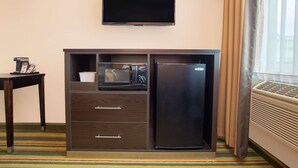 Habitación de hotel con televisión de pantalla plana en la pared, minibar, refrigerador, una mesita con una revista y ventana con cortinas.