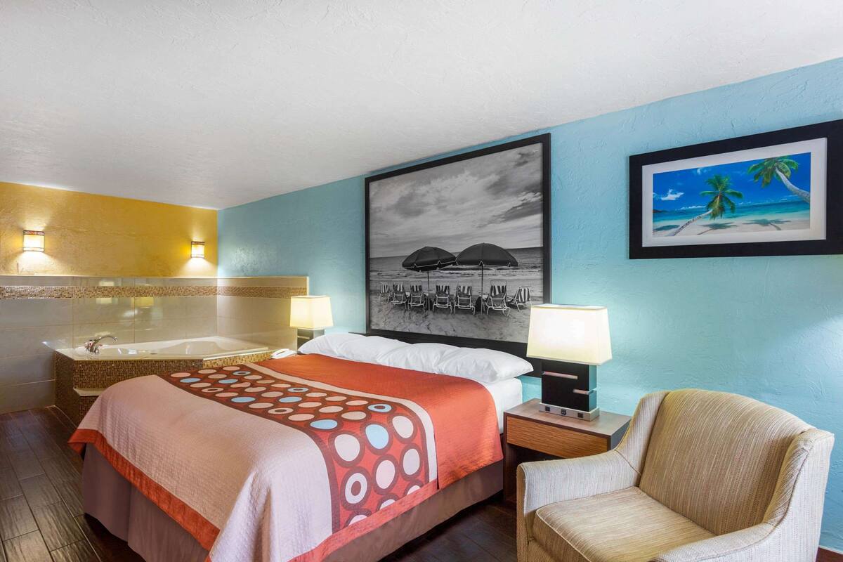 A hotel room with a bed, a chair, a large painting of a beach scene, a bathtub, and a wall-mounted lamp.