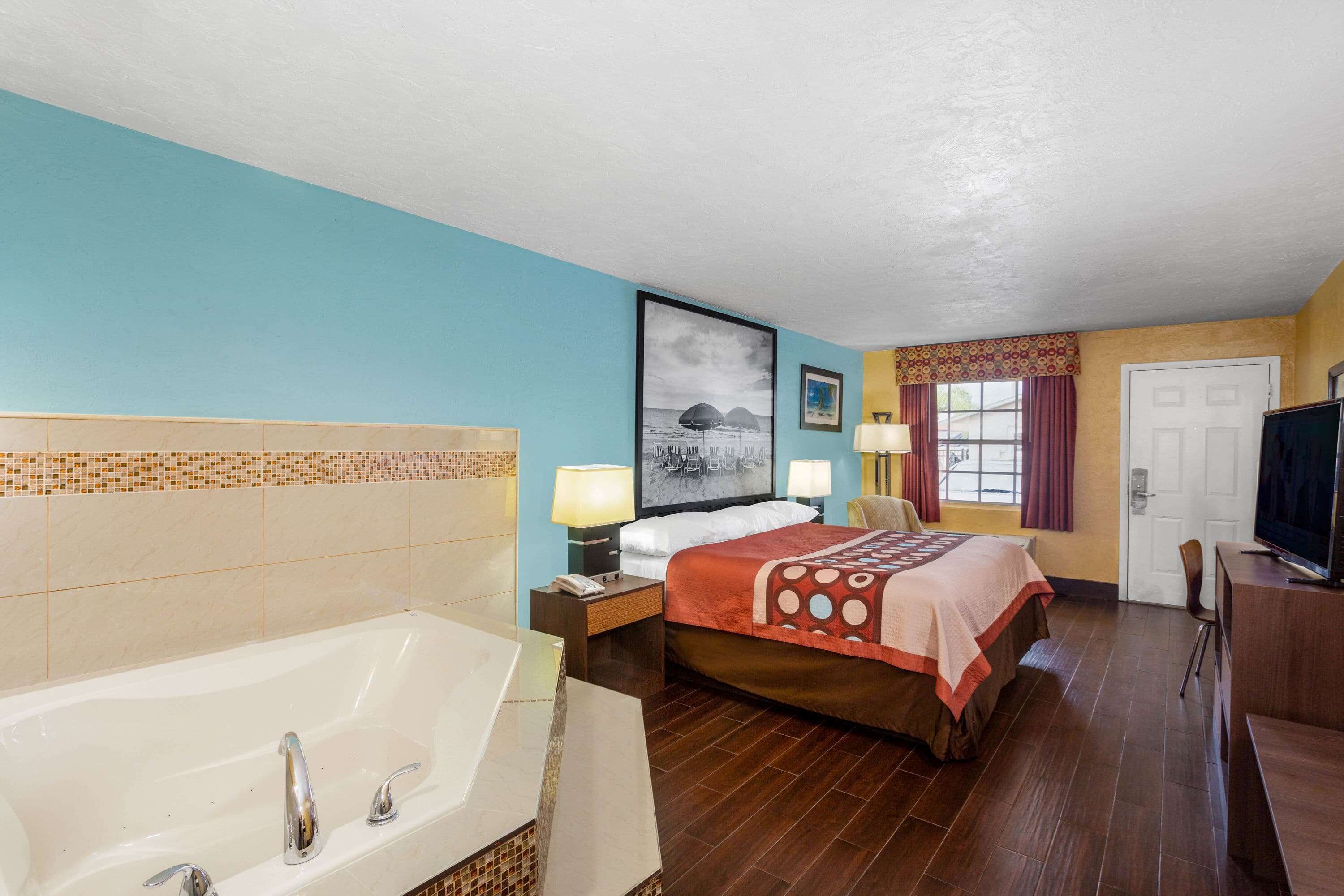 A hotel room with a large bathtub, a bed with a red and white patterned bedspread, two bedside lamps, a framed picture on the wall, and a television on a wooden stand.
