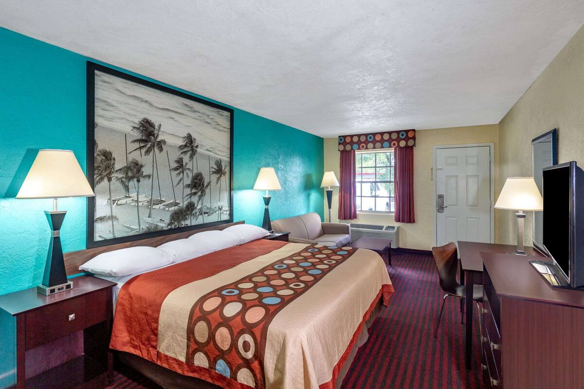 A hotel room with a large bed, two bedside tables with lamps, a desk with a chair, a TV, and a large painting on the wall.