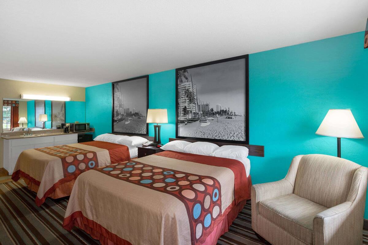 A hotel room with two beds, a chair, a desk with a lamp, and artwork on the walls.