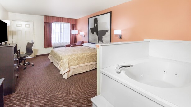 A hotel room with a large bed, a desk with a chair, a TV, a desk lamp, a large window with curtains, a wall-mounted artwork, and a bathtub.