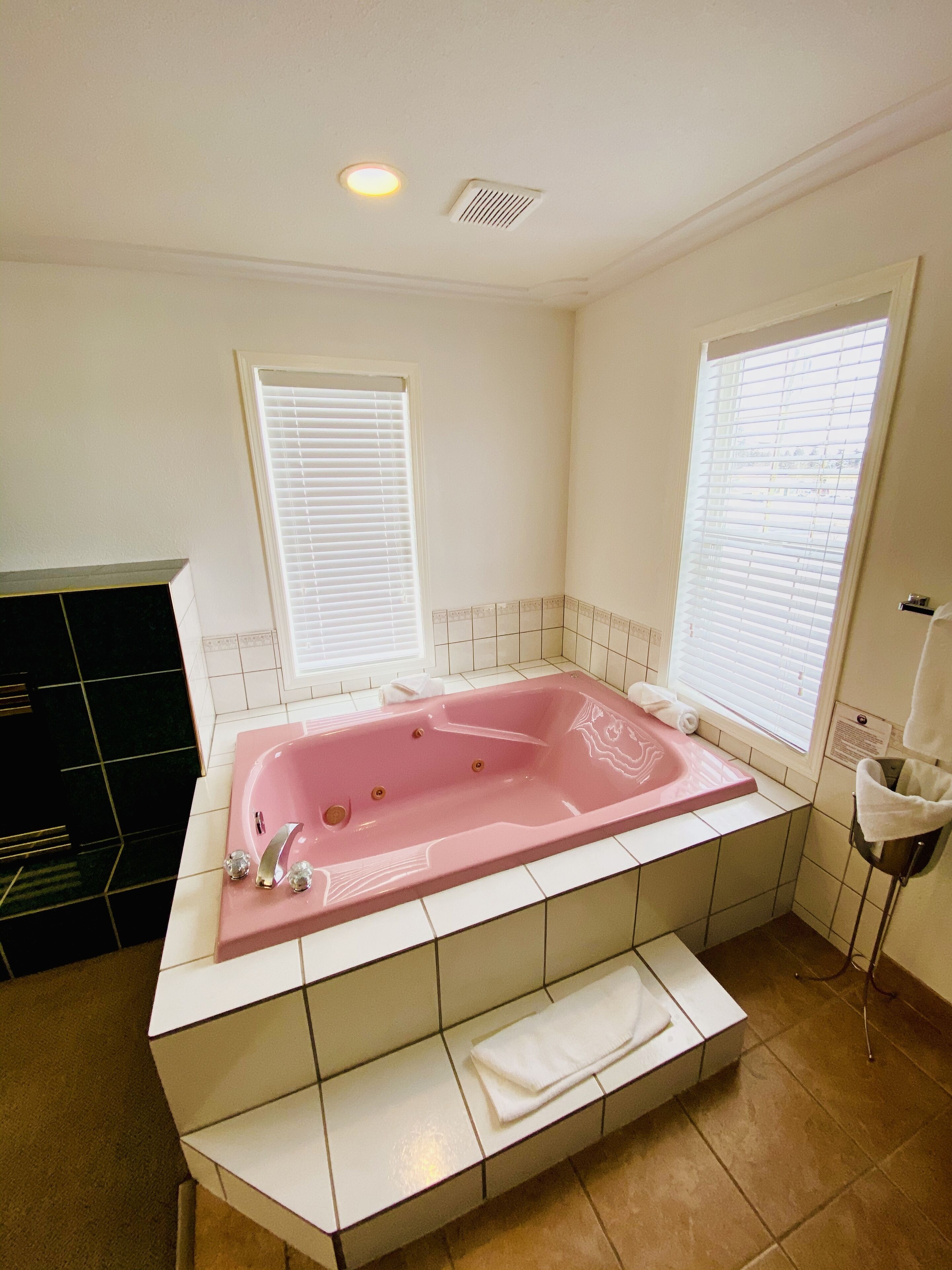 A bathroom with a large pink bathtub, white tiled walls, and two windows with blinds.