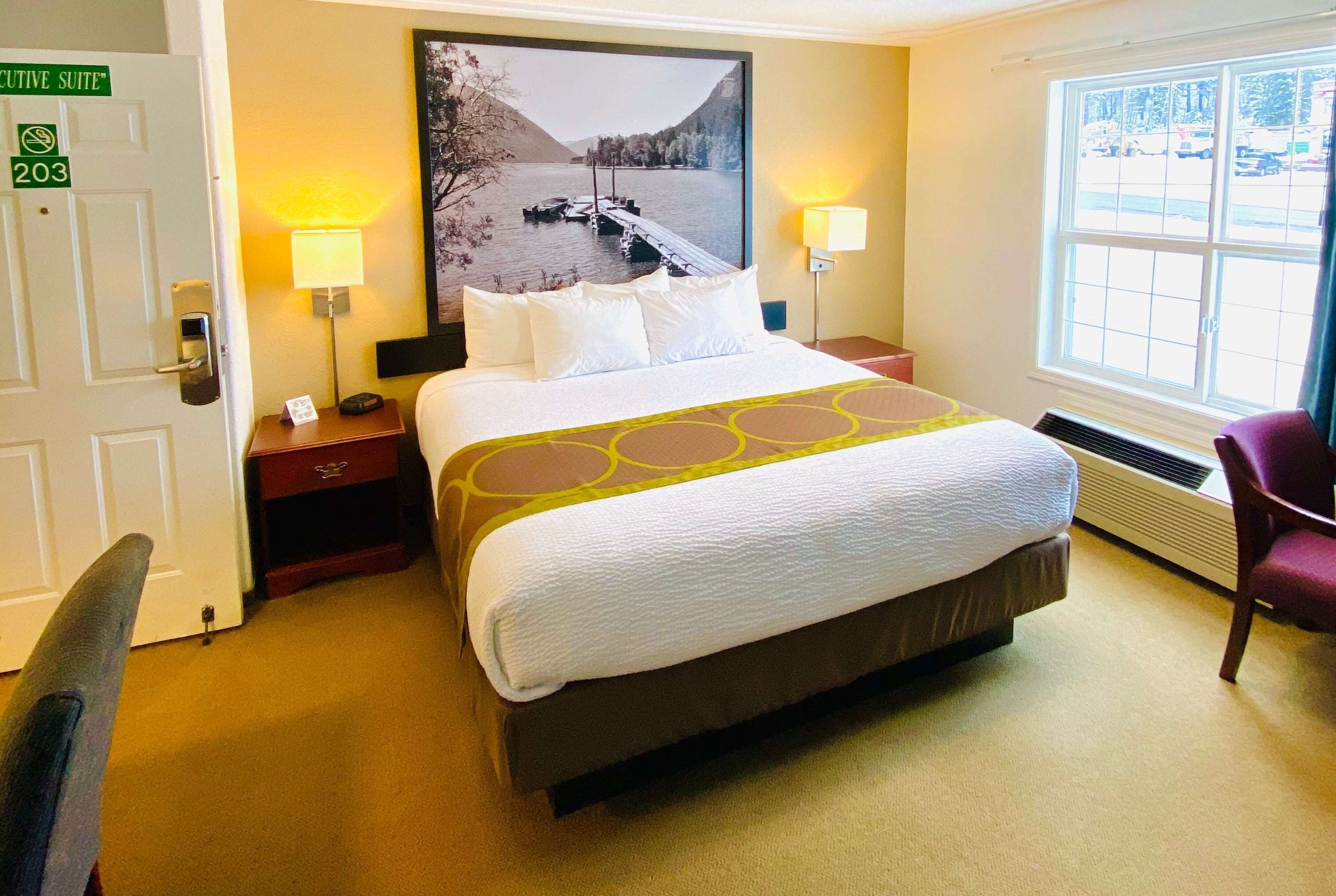 A hotel room with a large bed, two bedside lamps, a nightstand, a chair, a window with a view of the outdoors, and a door labeled "Executive Suite 203".