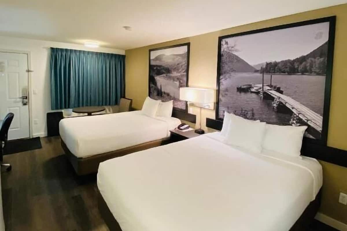 Habitación de hotel con dos camas, un escritorio, una silla, una lámpara y un gran ventanal con vistas a un lago y montañas.