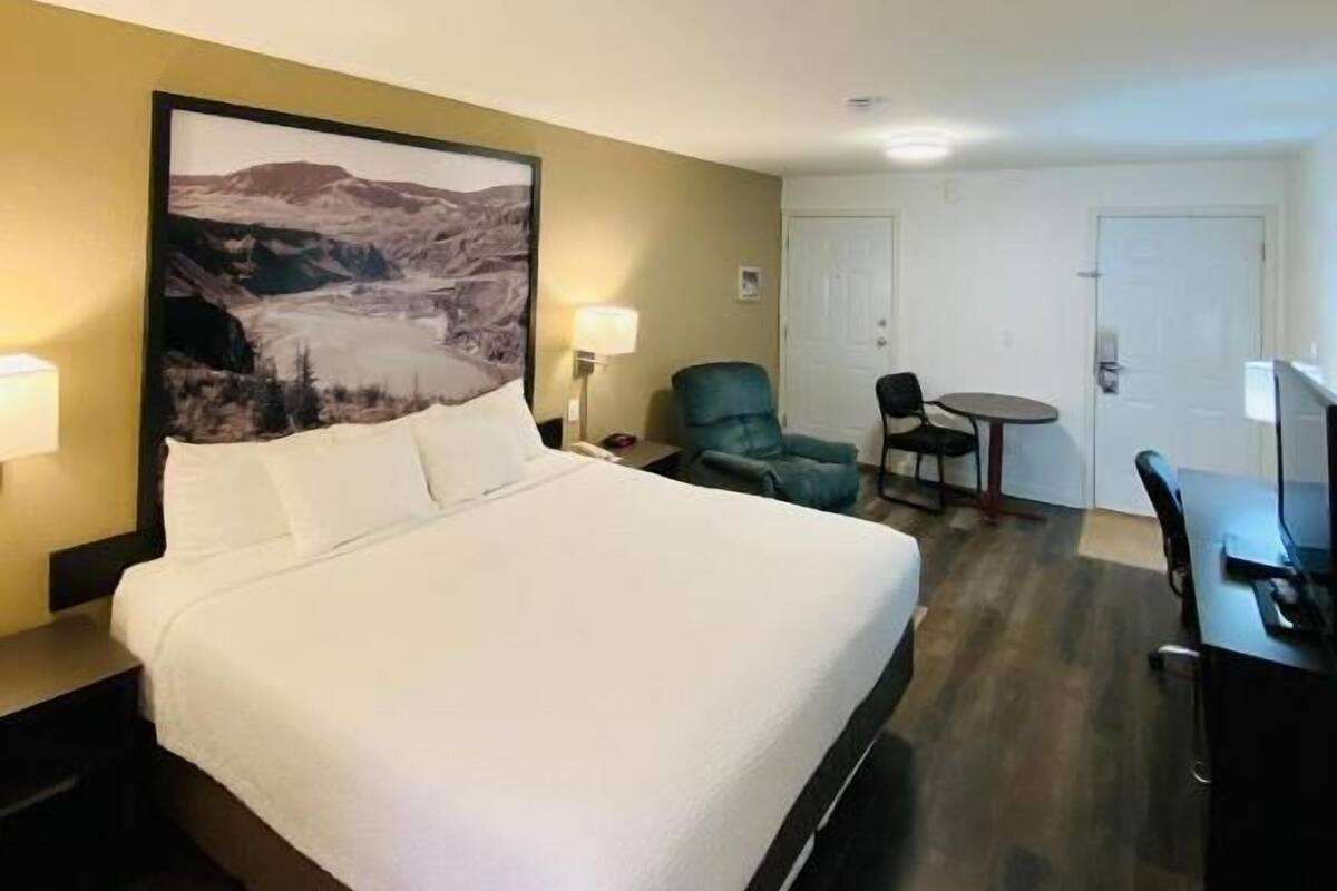A hotel room with a large bed, a desk with a chair, a small table, and a chair. There is a painting on the wall and a door in the background.