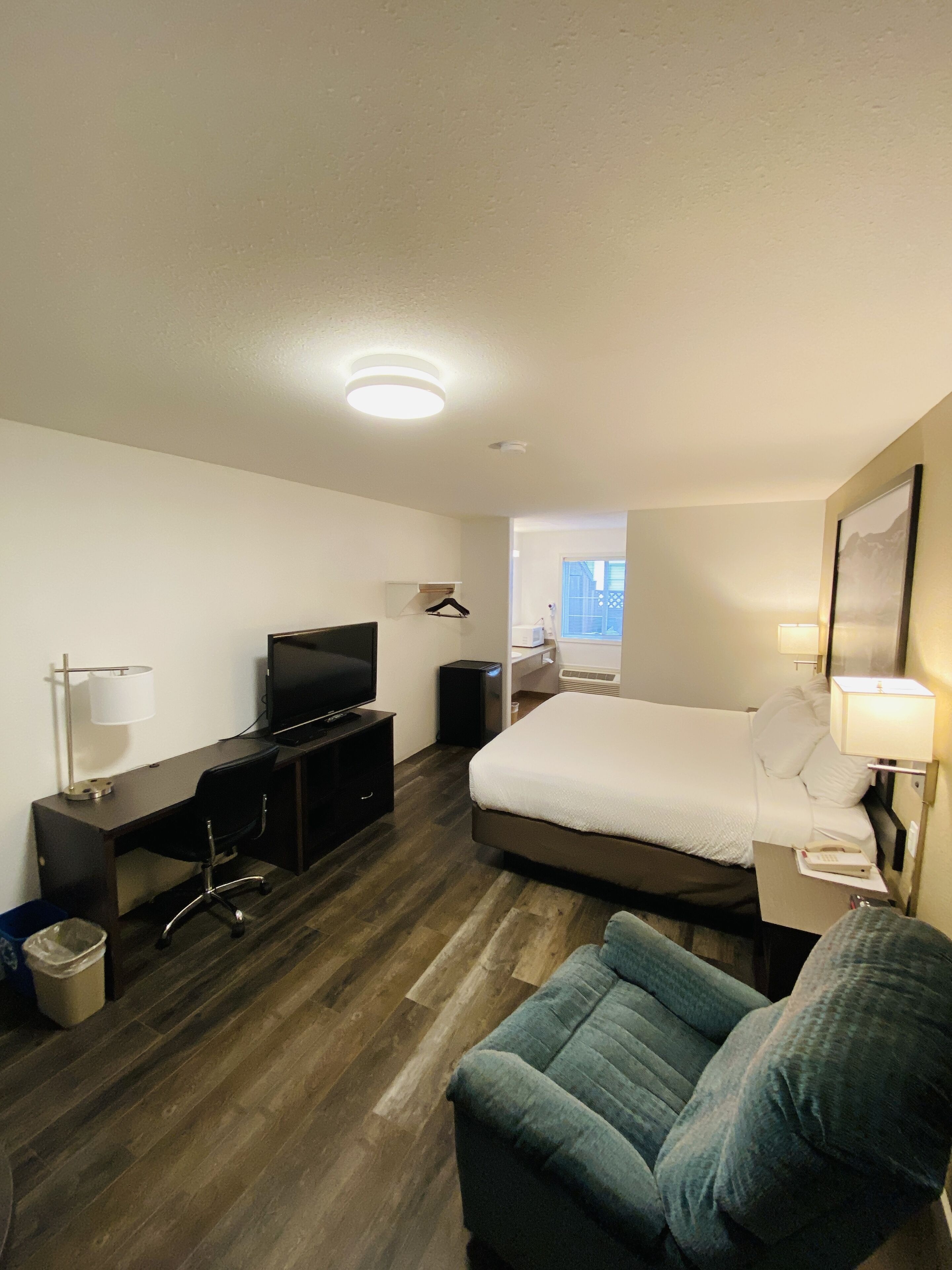 Ein Hotelzimmer mit einem Bett, einem Schreibtisch mit Fernseher, einem Sessel und einem kleinen Tisch mit Lampe.