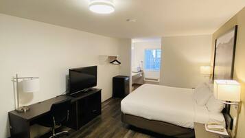 Ein Hotelzimmer mit einem Bett, einem Schreibtisch mit Fernseher, einem Sessel und einem kleinen Tisch mit Lampe.