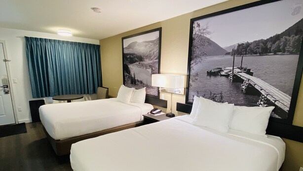 A hotel room with two beds, a desk, a chair, a lamp, and a large window with a view of a lake and mountains.