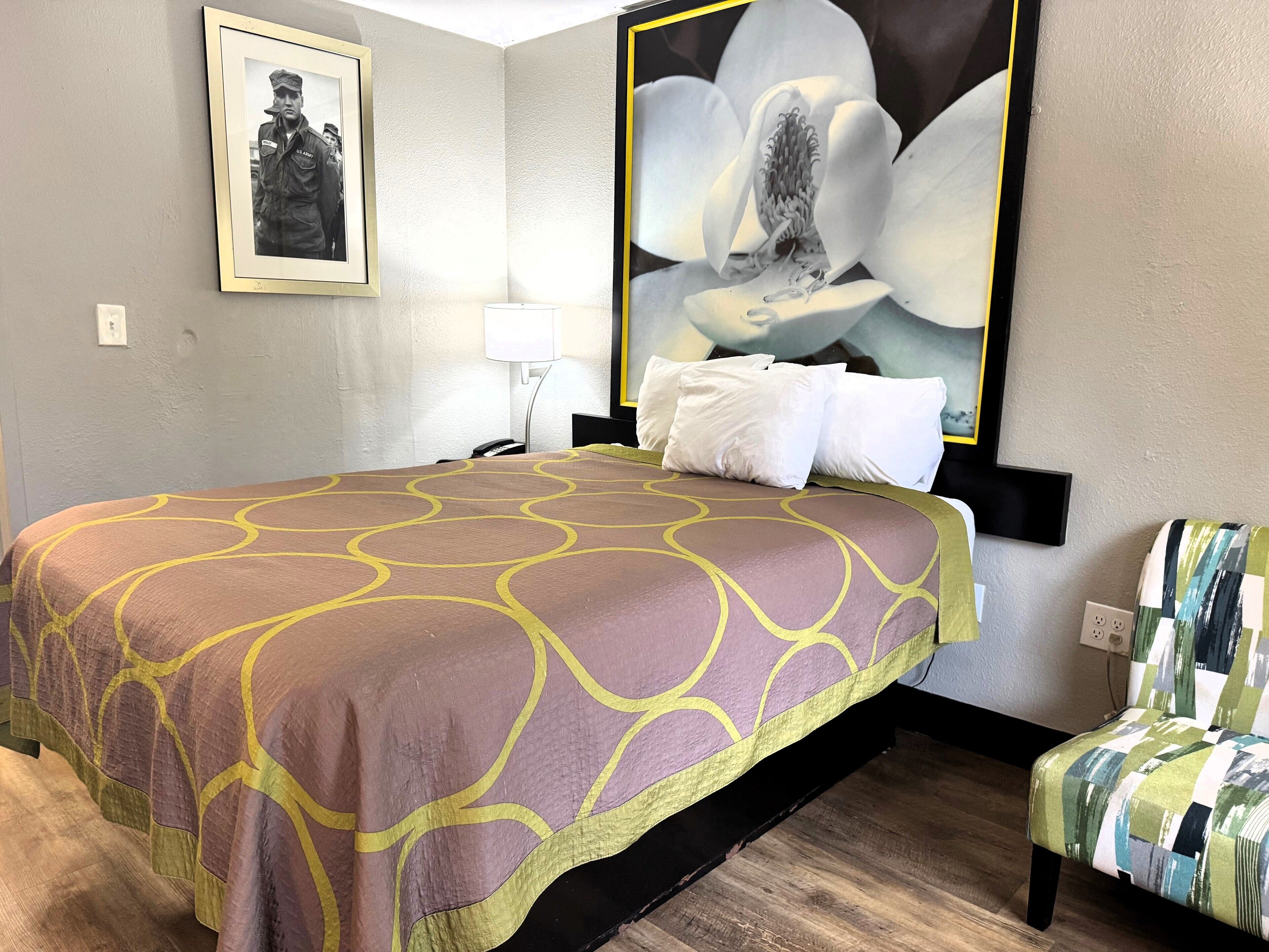 A hotel room with a bed, a nightstand, a lamp, a framed picture on the wall, and a large floral artwork.