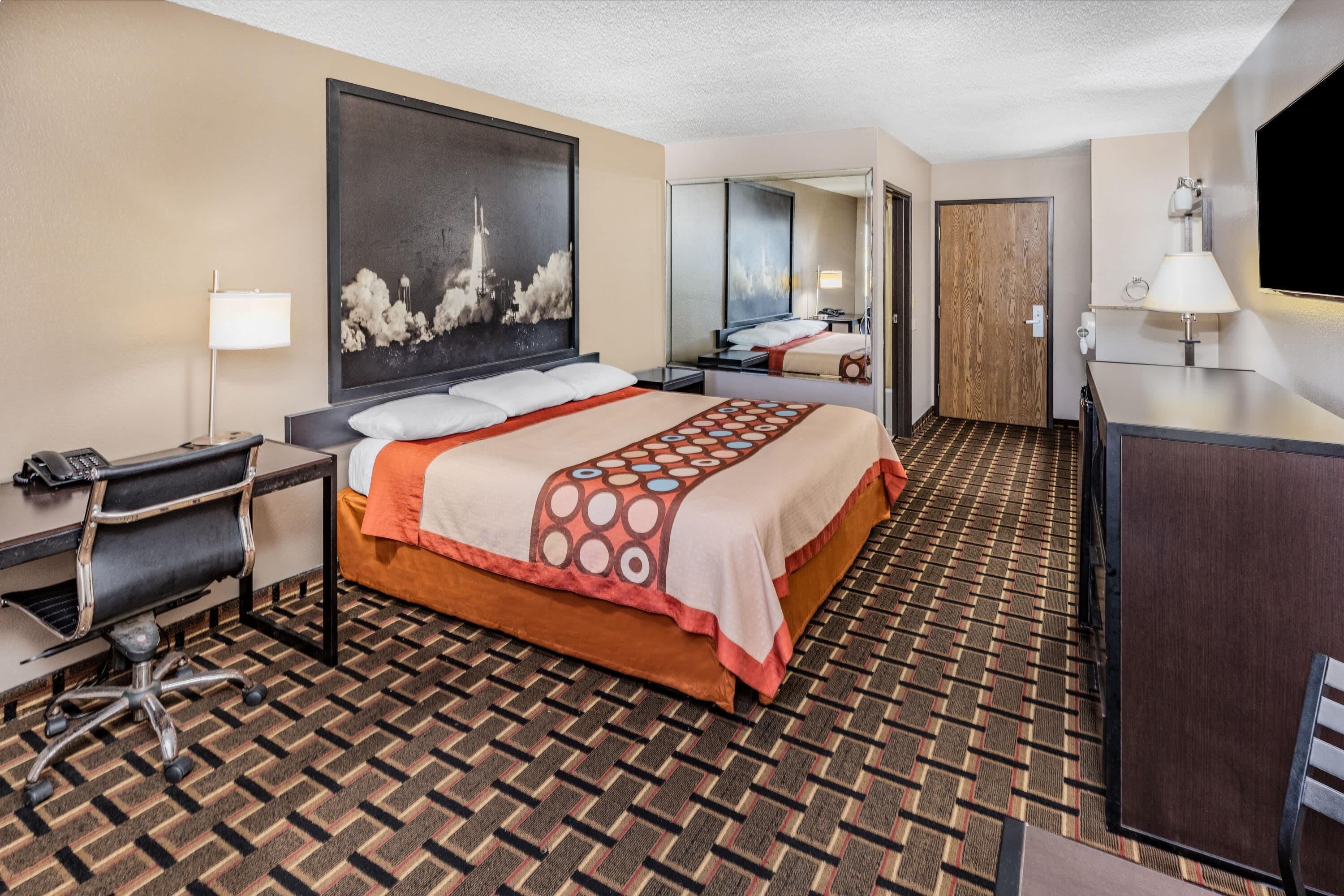 Quarto de hotel com cama, escrivaninha, cadeira e um espelho grande.