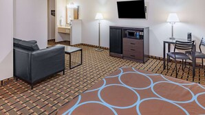 A hotel room with a sofa, a small table, a TV, a desk with a chair, and a minibar.