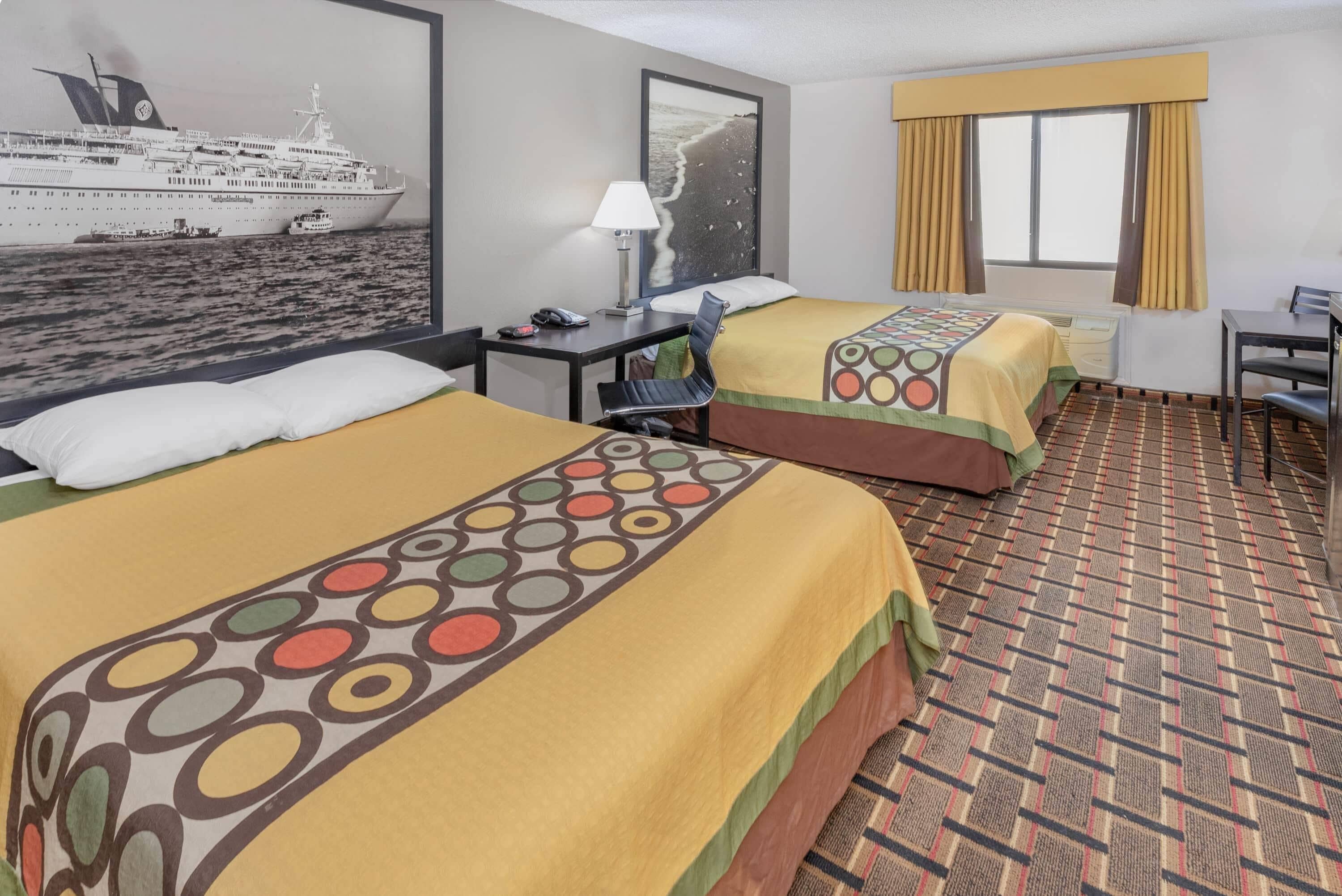 A hotel room with two beds, a desk, a chair, a window with curtains, and a wall-mounted lamp.