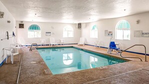 An indoor swimming pool with clear water, surrounded by white chairs and a blue chair, with arched windows and a ceiling with recessed lighting.