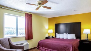 A hotel room with a bed, a chair, a ceiling fan, a window with curtains, and a wall-mounted heater.