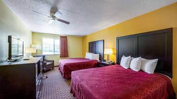A hotel room with two beds, a desk, a chair, a television, and a ceiling fan.