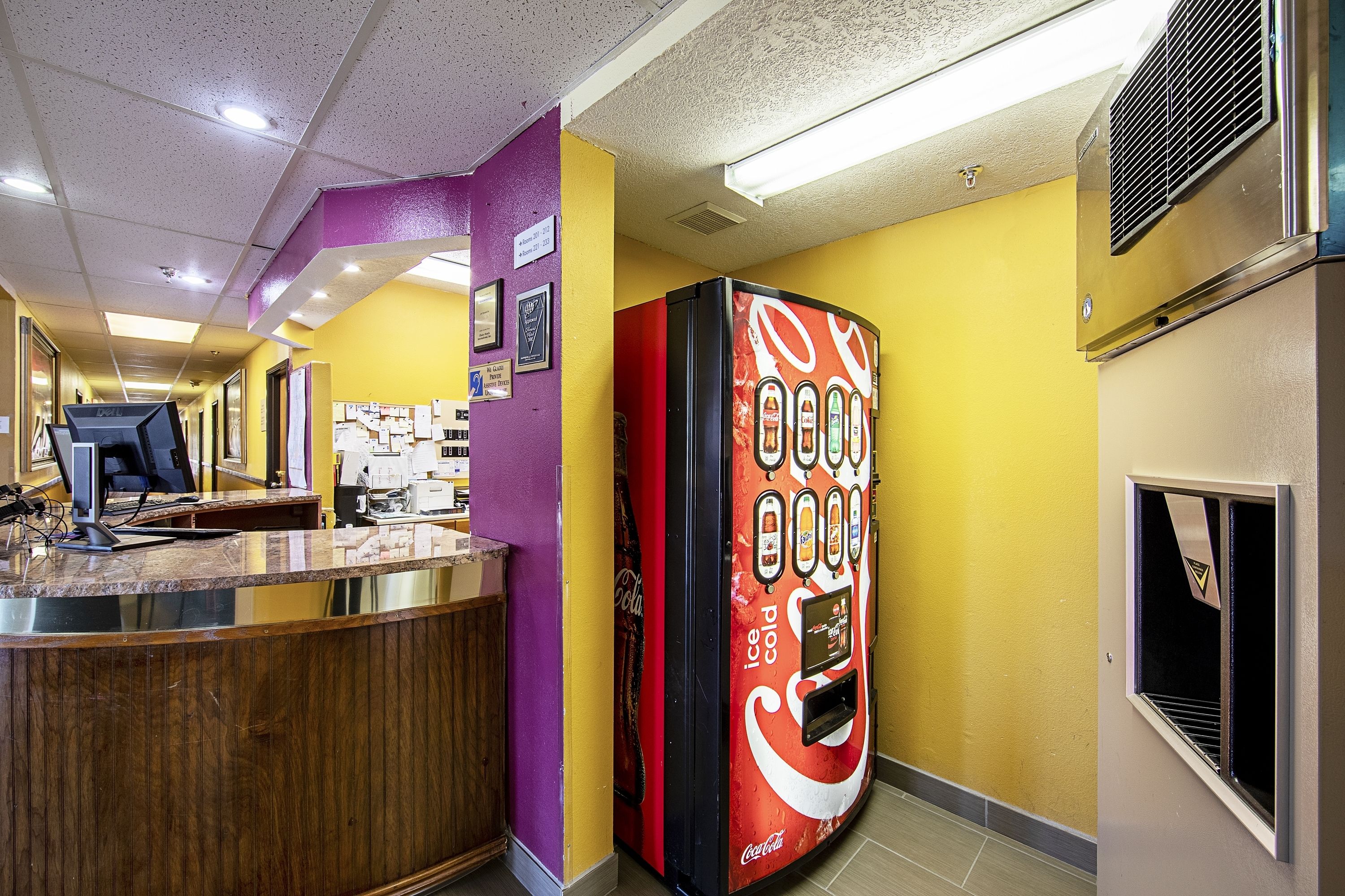 A hotel lobby with a reception desk, a Coca-Cola branded vending machine, and a purple wall with framed pictures.