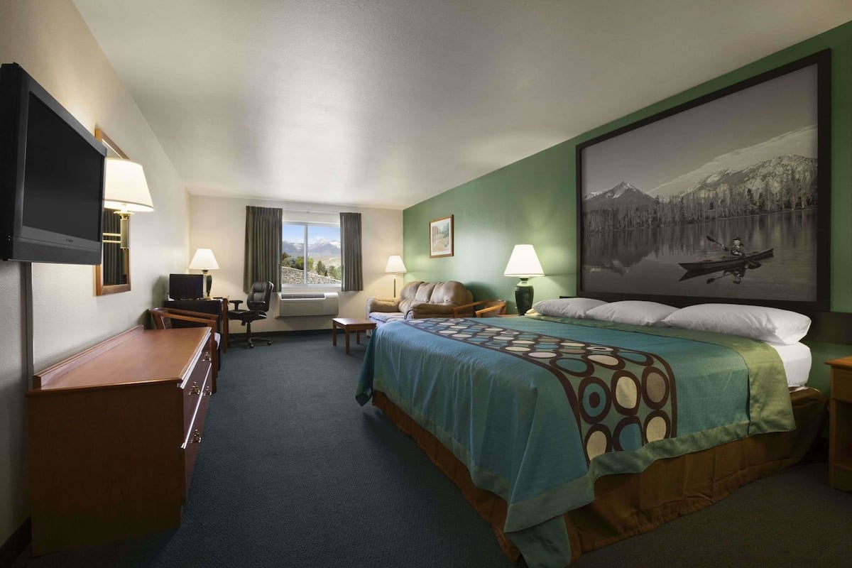 Quarto de hotel com cama, escrivaninha, cadeira, televisão, janela com cortinas e um quadro na parede.