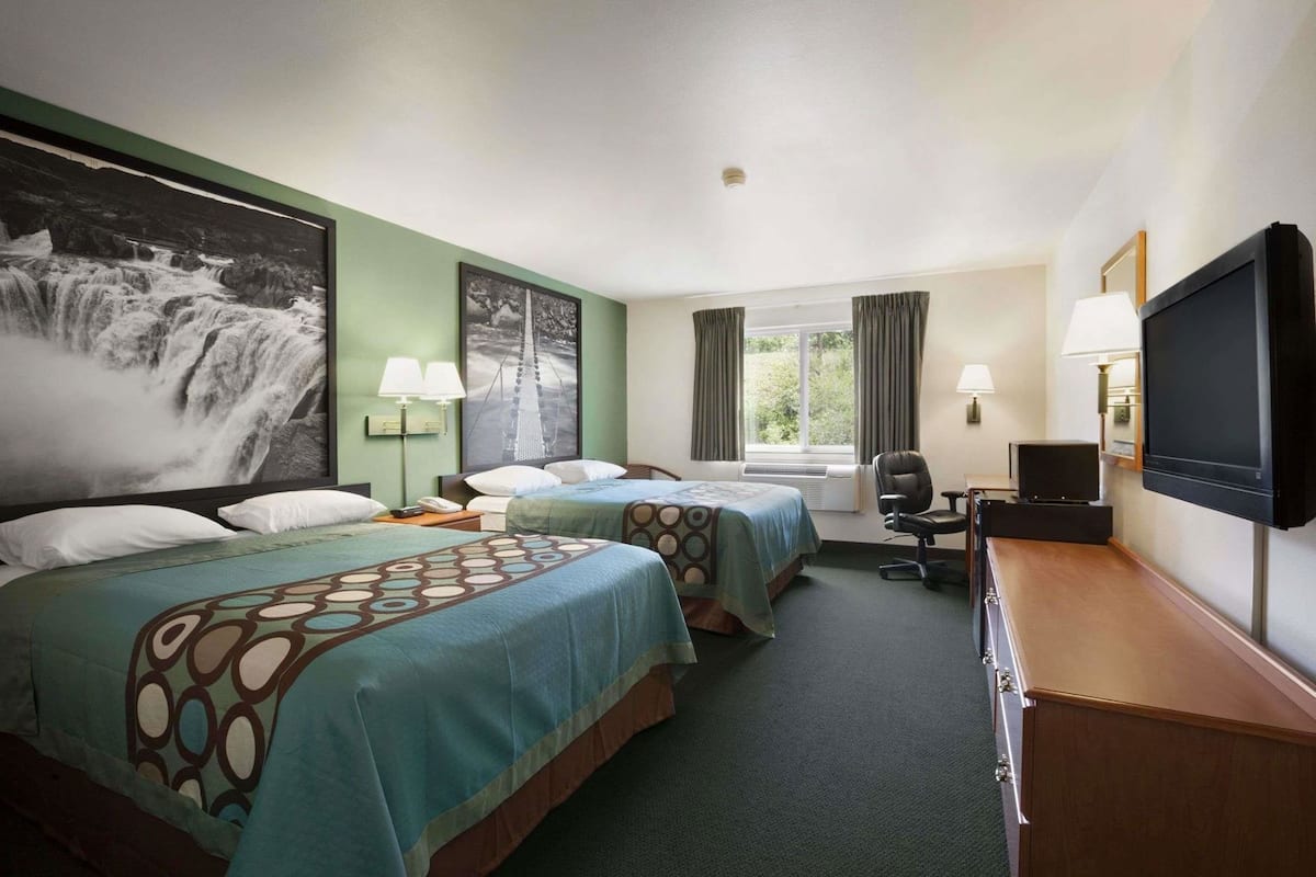 Quarto de hotel com duas camas, uma mesa com televisão, uma cadeira e uma janela grande com cortinas.