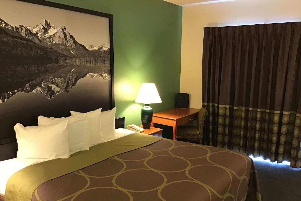 Quarto de hotel com cama, escrivaninha, luminária, telefone e janela com cortinas.