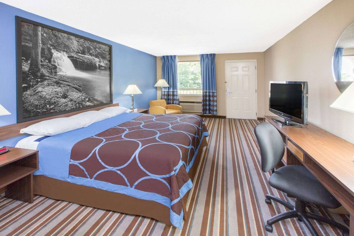 A hotel room with a bed, a desk with a TV, a chair, a lamp, and a large framed picture on the wall.