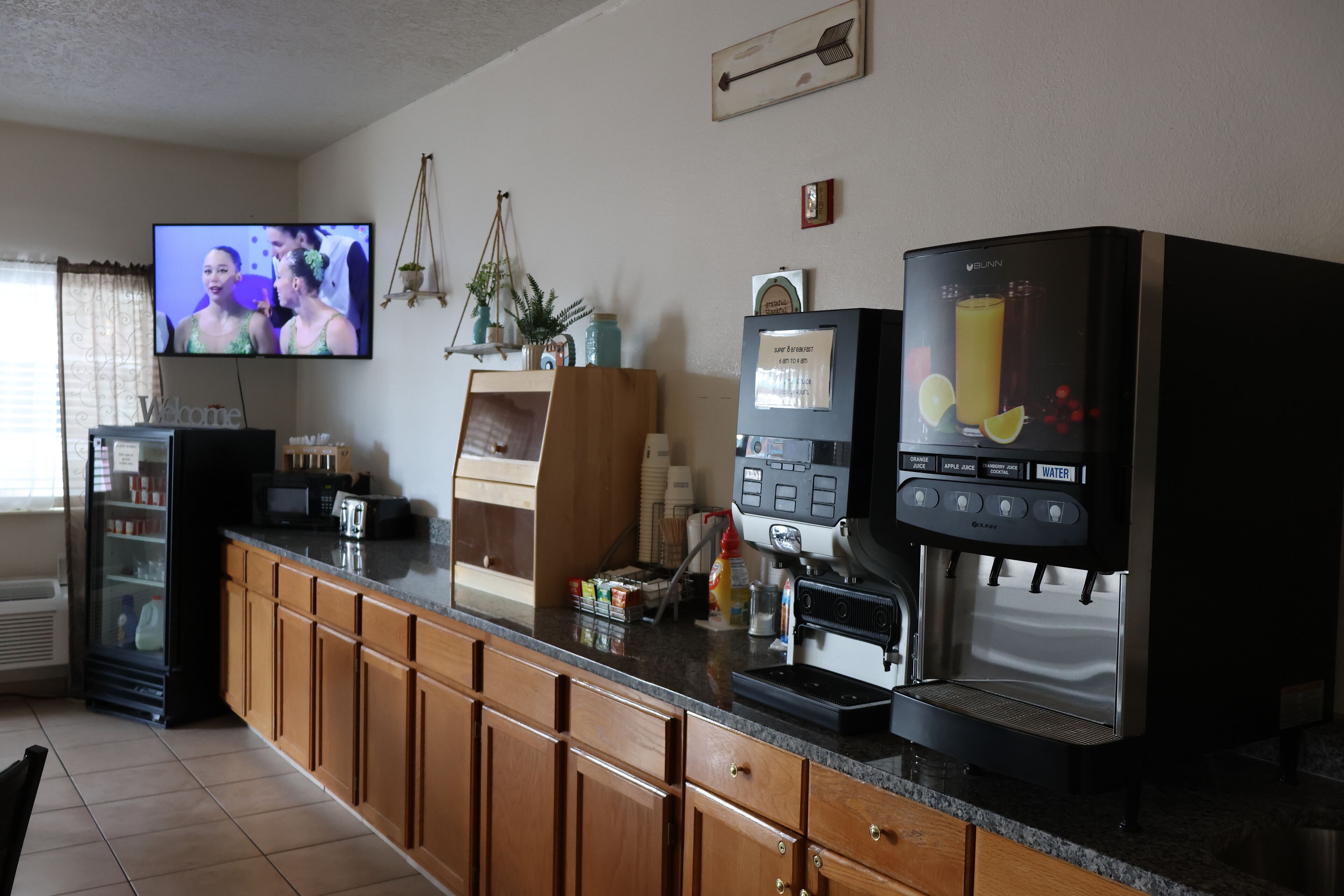 Une cuisine avec des meubles de rangement en bois, un réfrigérateur, un four à micro-ondes, une machine à café et un téléviseur qui diffuse une scène.