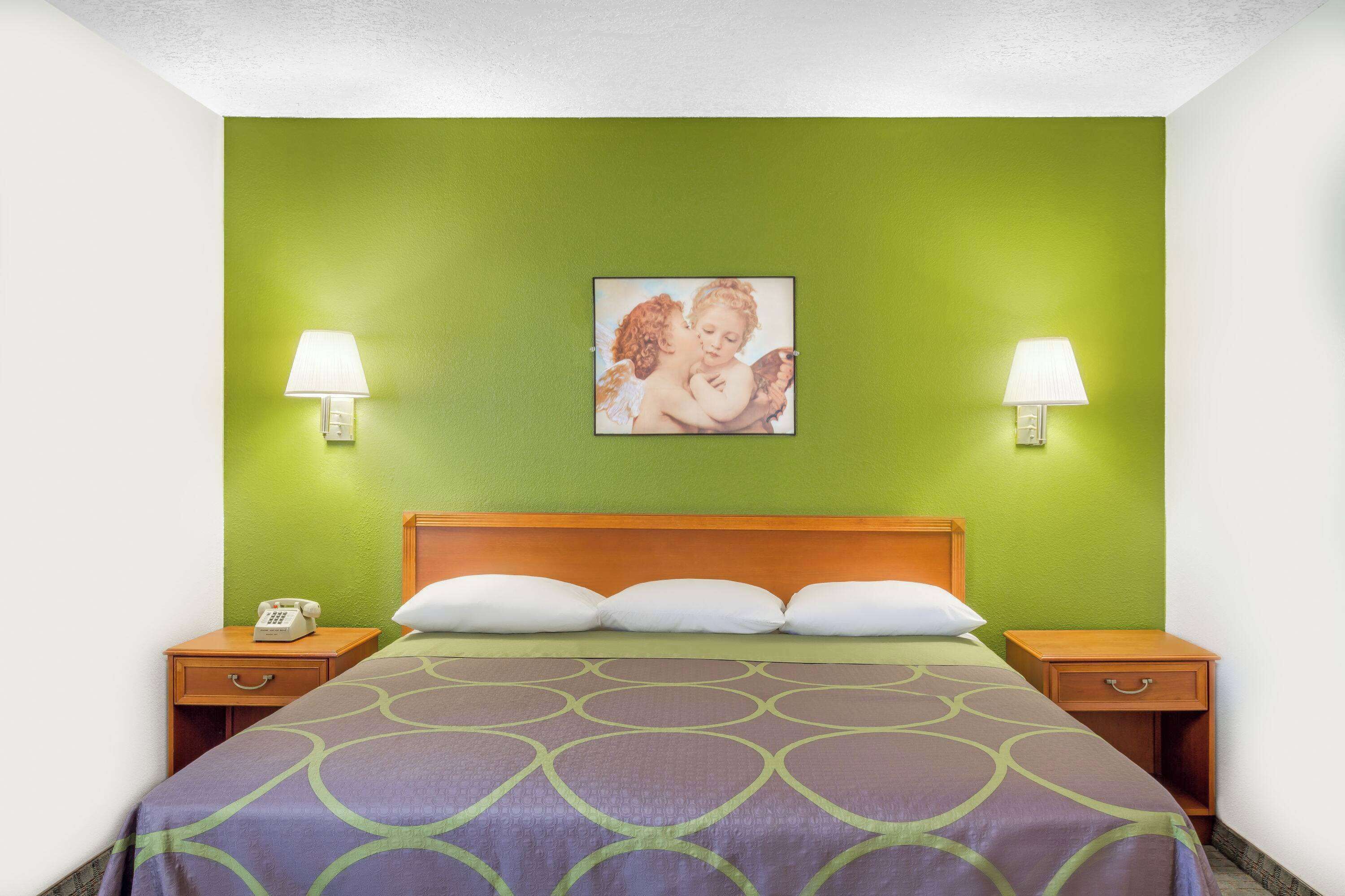 Habitación de hotel con pared verde, cabecera de madera, cama con colcha estampada, dos mesitas de noche con lámparas y un cuadro enmarcado en la pared.