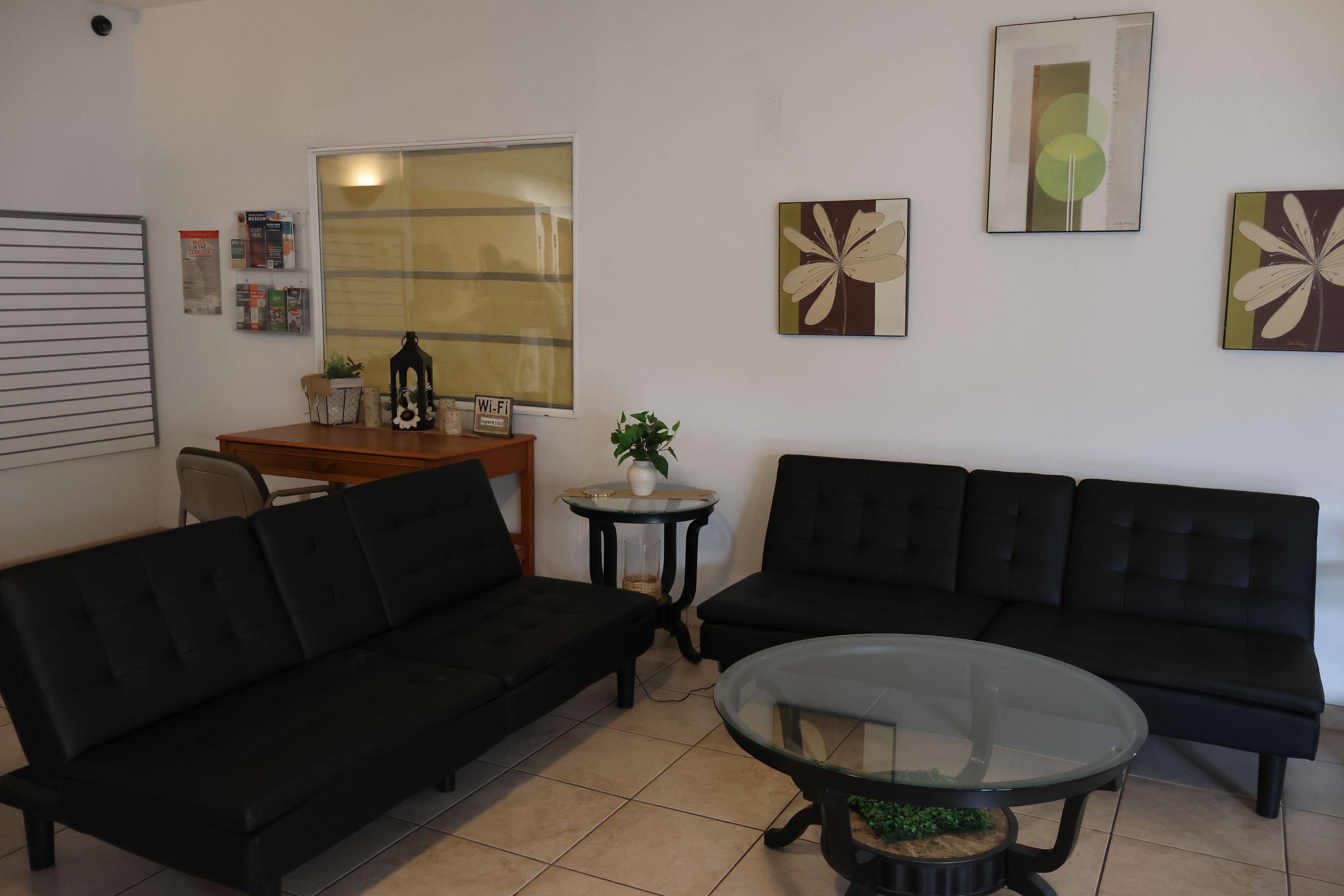 Un salón con dos sofás negros, una mesa de centro de cristal, una mesita auxiliar con una planta y una ventana con persianas.