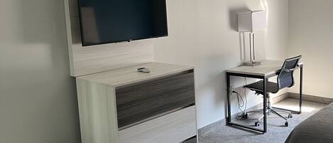 A modern hotel room with a flat-screen TV mounted on a wall, a desk with a chair, a bedside table, and a bed.