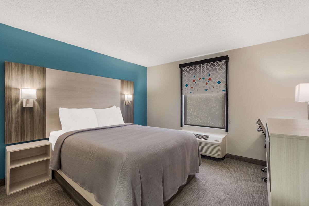 A hotel room with a bed, bedside tables, a nightstand, a window with a geometric pattern, and a wall-mounted air conditioning unit.