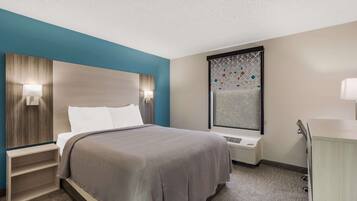 A hotel room with a bed, bedside tables, a nightstand, a window with a geometric pattern, and a wall-mounted air conditioning unit.
