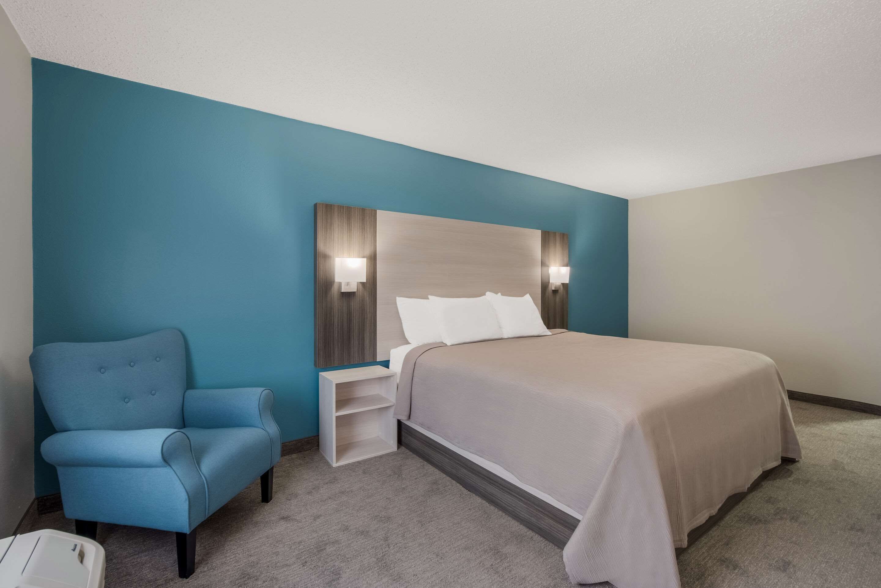 A hotel room with a bed, a nightstand, a chair, and a wall-mounted lamp.