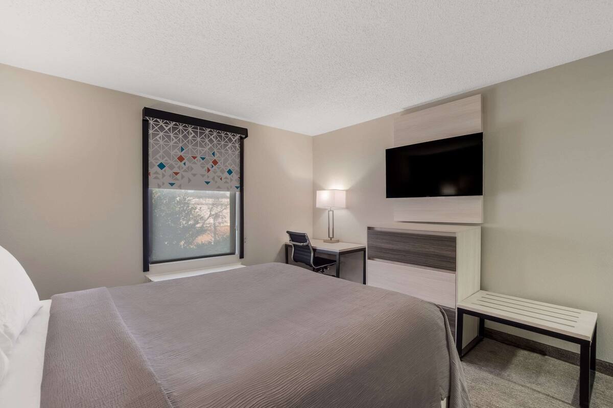 A hotel room with a large bed, a desk with a chair, a TV mounted on the wall, and a window with patterned curtains.