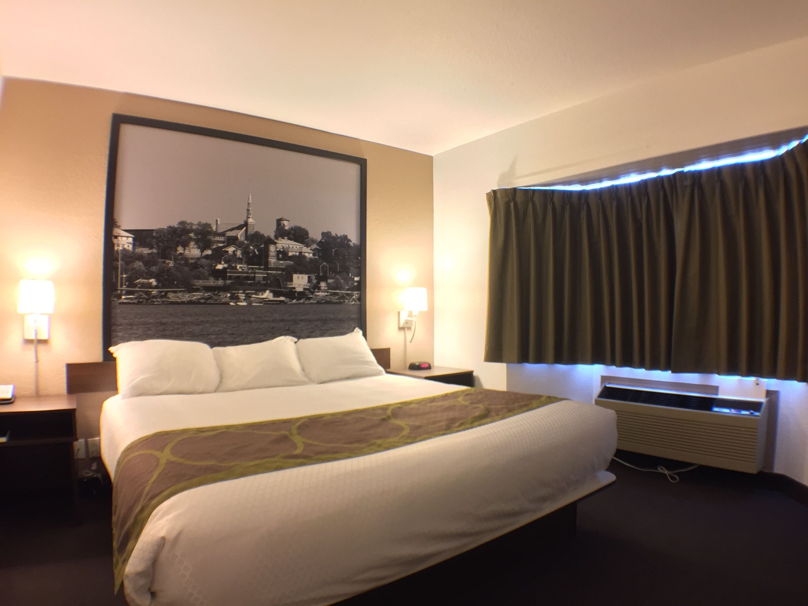 Une chambre d’hôtel avec un grand lit, deux lampes de chevet, un tableau représentant un paysage urbain accroché au mur, une fenêtre avec des rideaux et un appareil de climatisation.