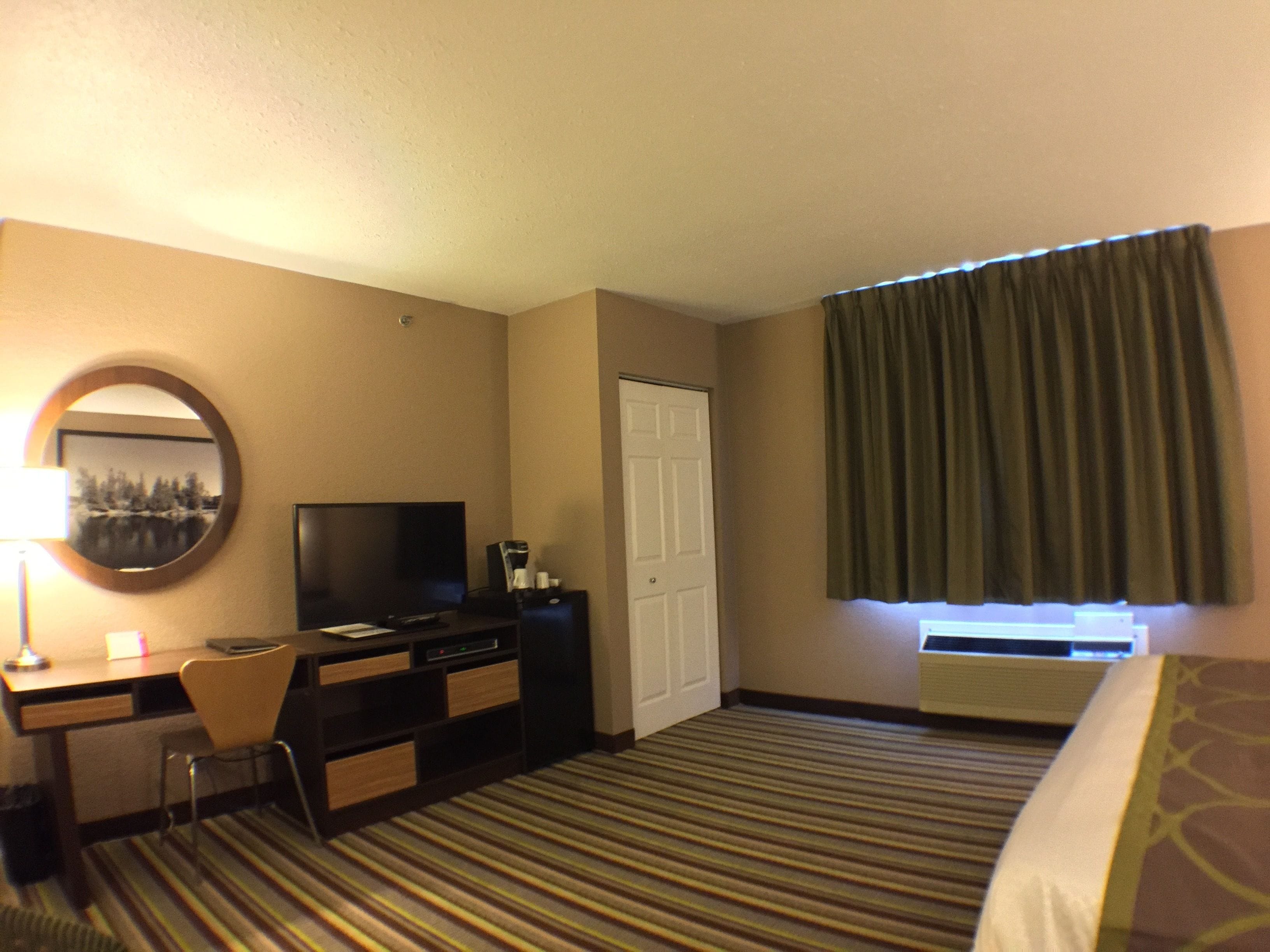 A hotel room with a bed, a desk with a chair, a television, a dresser, a mirror, and a window with curtains.